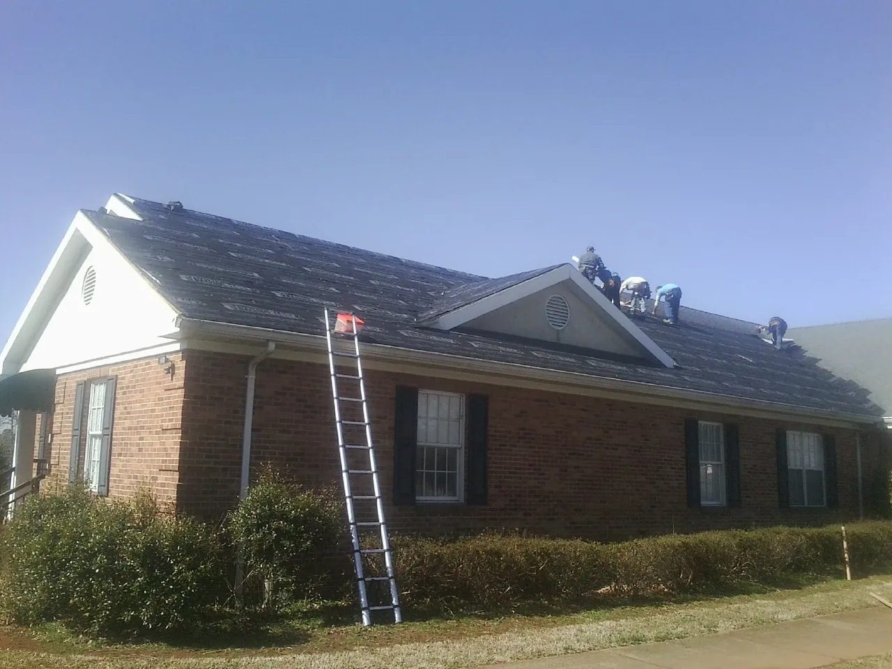 Crew tearing shingles off a brick ranch roof with a ladder set — Metal Roof Repair work by our crew in Ionia